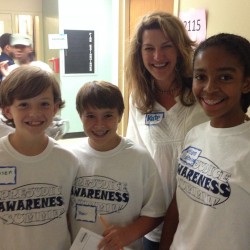 A group of three young students in matching shirts are smiling with a woman standing behind them, smiling as well.