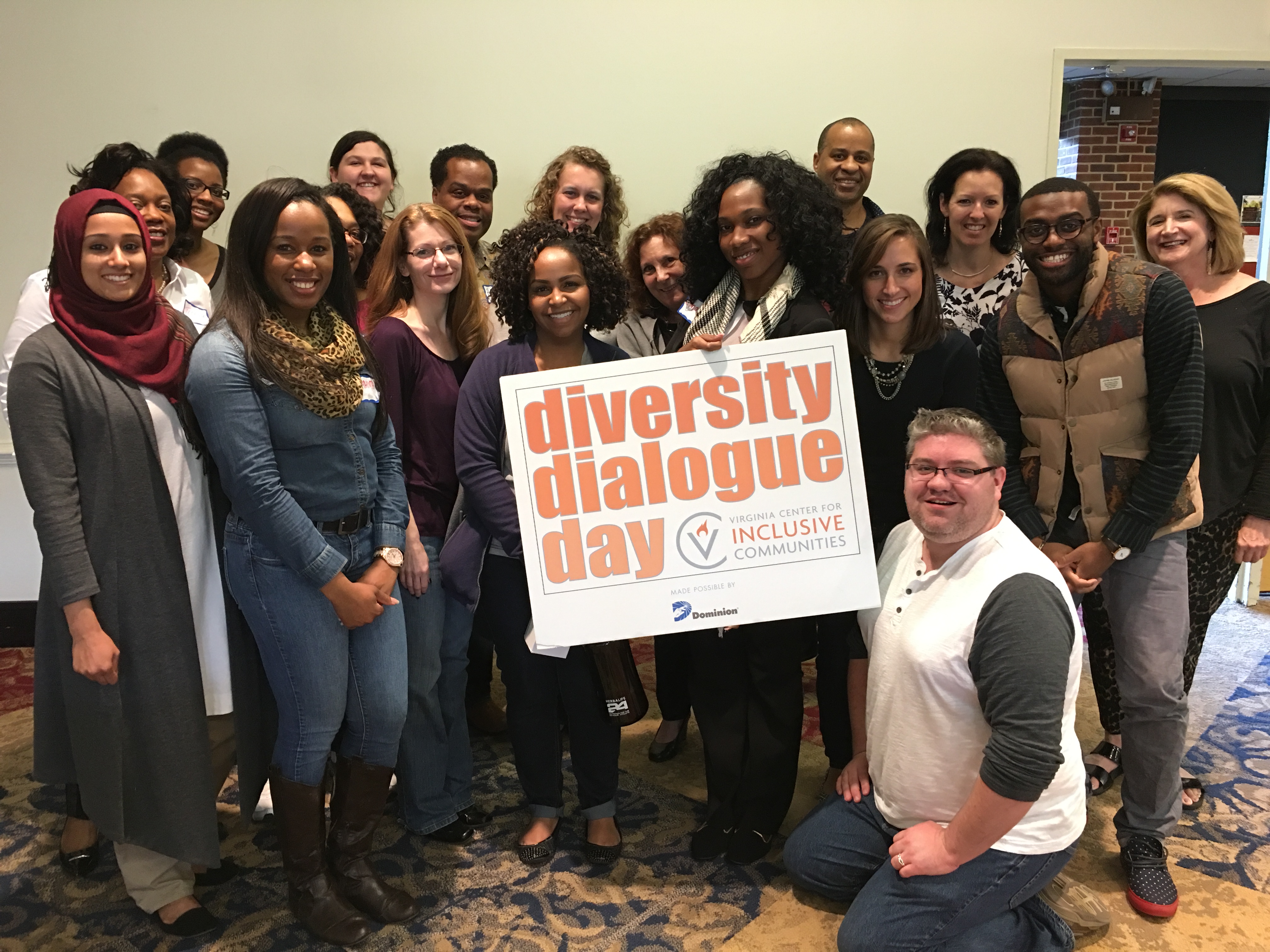 A diverse crowd of people are gathered together. They are smiling and posing while holding up a sign reading “diversity dialogue day”.