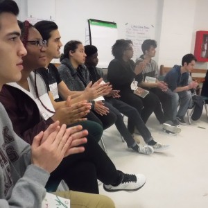 A group of eight students participating in a workshop in a classroom applaud.