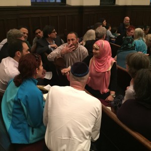A gathering of at least twenty people is taking place over five rows of pews. People are engaging in conversation with those around them.