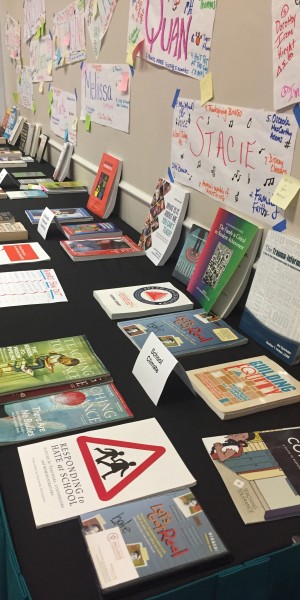 A table with an array of books and pamphlets are categorized by different themes that are listed on tags. On the wall behind the table, colorful and decorated newsprints are taped on the wall with people’s names written on them. Several post-it notes are on each newsprint.