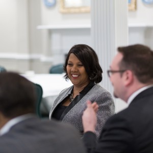 Three individuals engaged in conversation with the focus on the women in the middle. There are two men with all three in business attire. The woman is smiling.