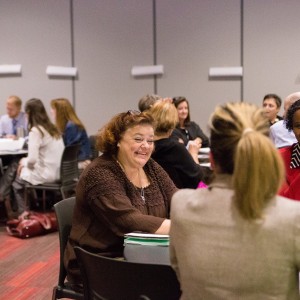 Groups of people in business attire are sitting at tables and talking with those at their table. The picture is focused on a woman smiling while engaging with those at her table.