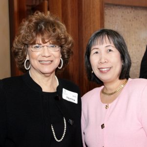 Two women smile at a camera. Both are dressed in business attire and one has a nametag on.