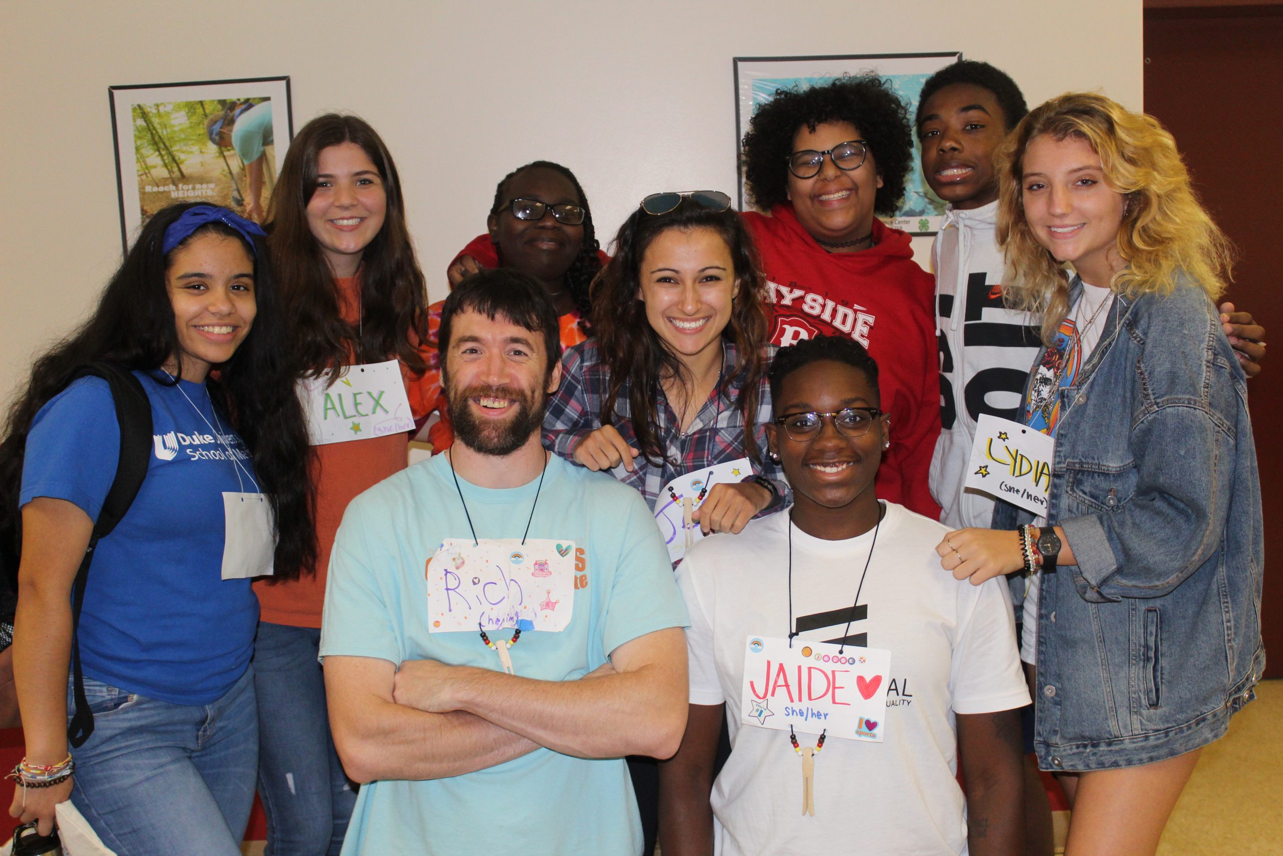 A diverse group of nine young people are standing together and smiling. They each have a paper nametag on that have been decorated and include the person’s name and pronouns.