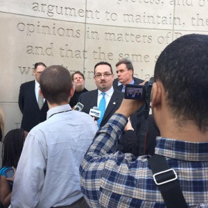 A man in business attire is speaking in front of a monument with other men in business attire behind him. There are several individuals holding microphones towards the man speaking and a person with a camera recording the man speaking.