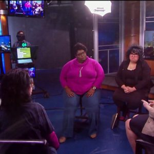 Four people are seen on a production set. There are teleprompters, television screens, and a light in the back. Four women are sitting with two slightly out of camera. The women are turned to the women in the center who is slightly out of view.