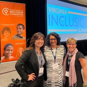 3 women stand together posing in front of a VCIC banner and a Virginia Inclusion Summit PowerPoint