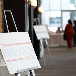 a white poster board with the Virginia Inclusion Summit logo is propped up on a metal stand. In the back ground, a blurry image shows 3 individuals around a table. 