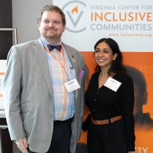 two Virginia Inclusion Summit attendees smile for a camera to the left, standing in front of a tall white and orange banner